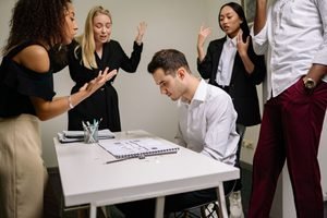 A man in a white shirt sits at a desk with his head bowed, looking down at an open notebook, while four colleagues stand around him gesturing with raised hands as if criticizing or reprimanding him.